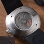 Stainless steel watch with exposed mechanical caseback showing orange Ikepod logo and black rubber strap on wooden surface.