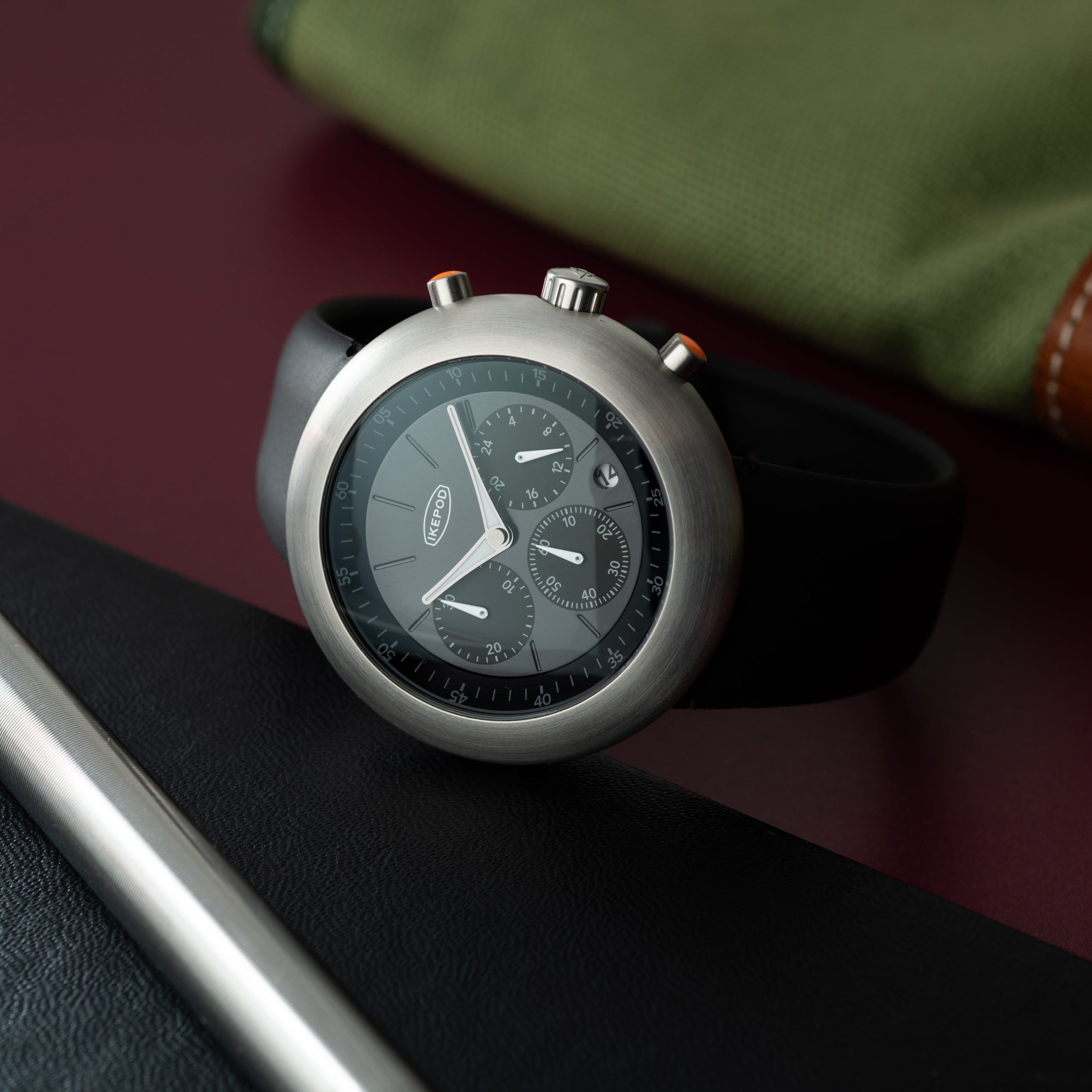 Black dial chronograph watch with silver case and black rubber strap on burgundy and green background.
