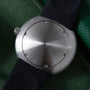 Brushed stainless steel watch face with black rubber strap and crown, displayed on green textured background.