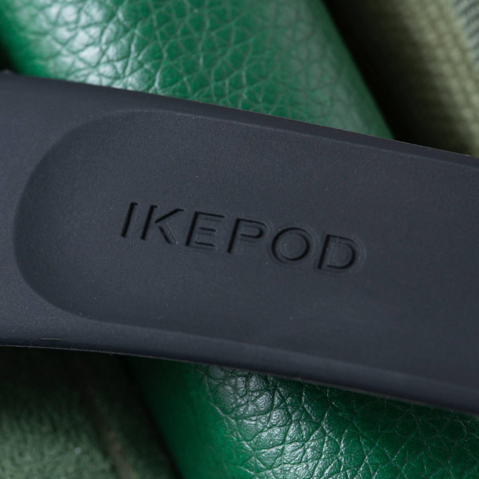 Black Ikepod watch case with embossed branding resting on green textured leather surface.