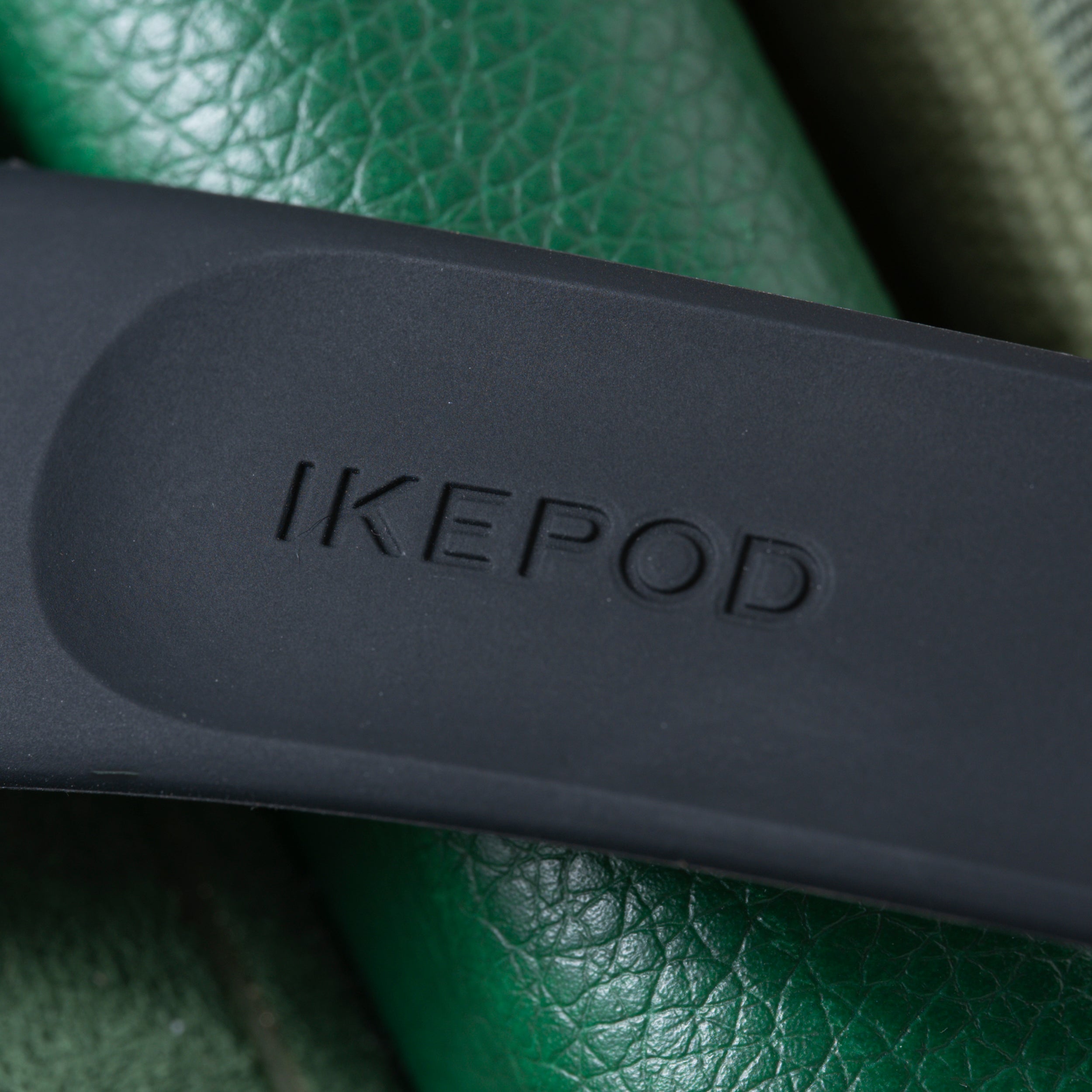 Black Ikepod watch case with embossed branding resting on green textured leather surface.