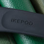 Black Ikepod watch case with embossed branding resting on green textured leather surface.