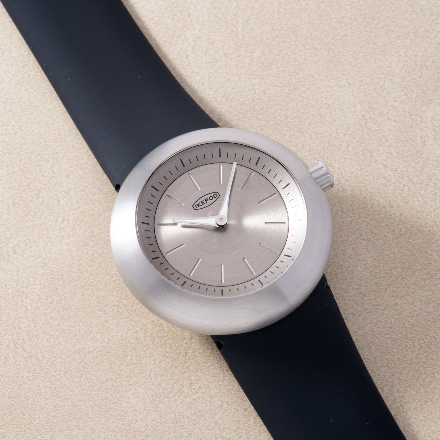 Round silver watch with gray dial, hour markers, and black rubber strap against beige background.