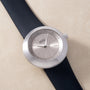 Round silver watch with gray dial, hour markers, and black rubber strap against beige background.
