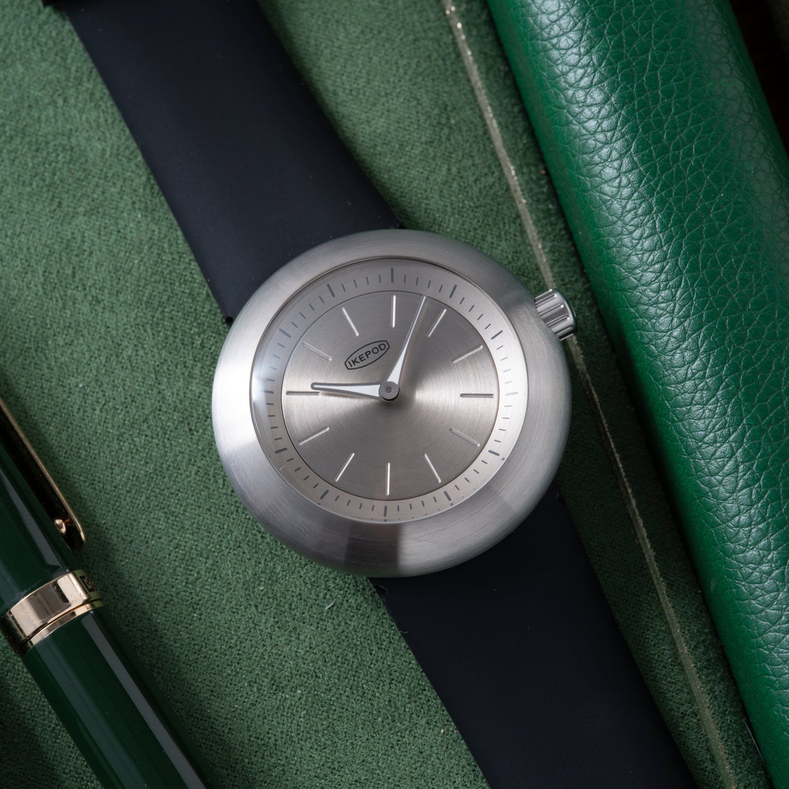 Ikepod Duopod watch with round brushed steel case, grey dial, and dark navy leather strap on green textile background.
