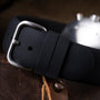 Black rubber strap with silver buckle and metal studs on brown suede surface.