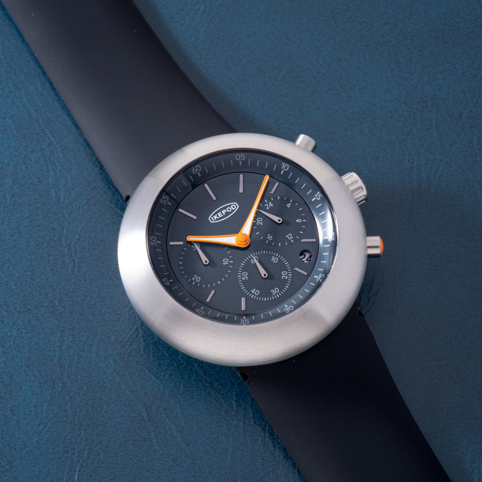 Silver-cased chronograph watch with dark dial, orange accents, and black rubber strap on blue background.