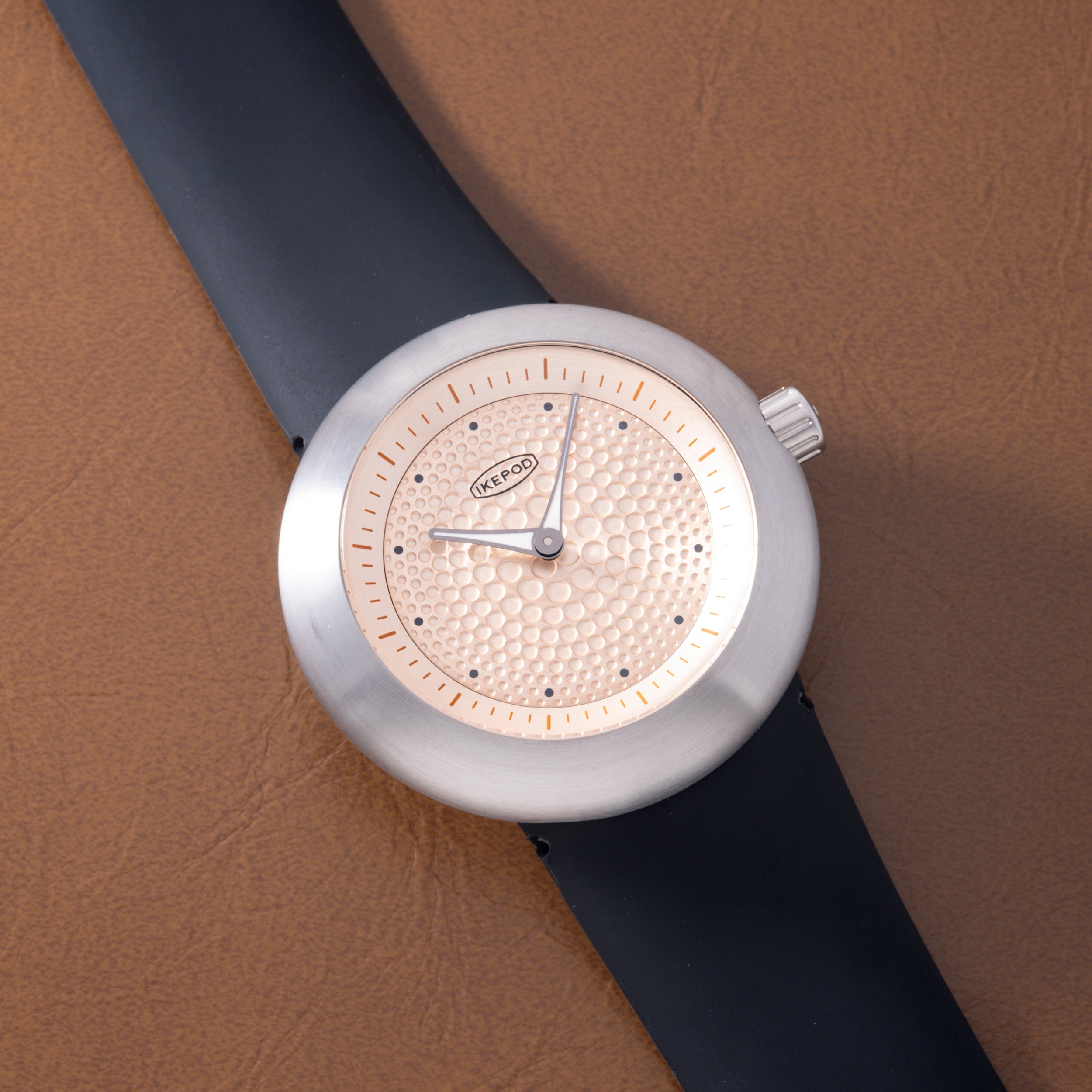 White round watch with textured gold dial, black leather strap, and hour markers on beige background.