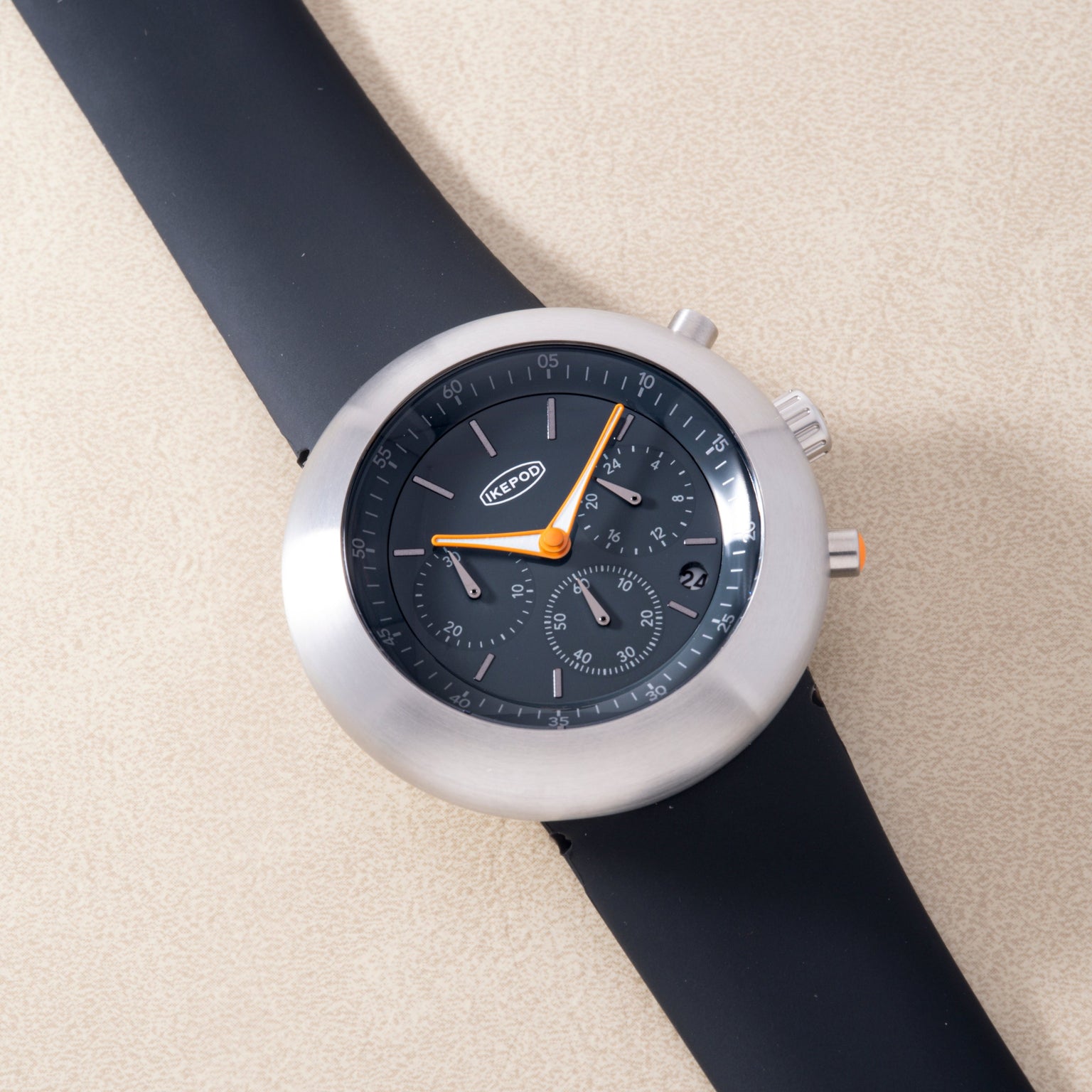 White chronograph watch with black dial, orange hands, and black rubber strap on beige background.