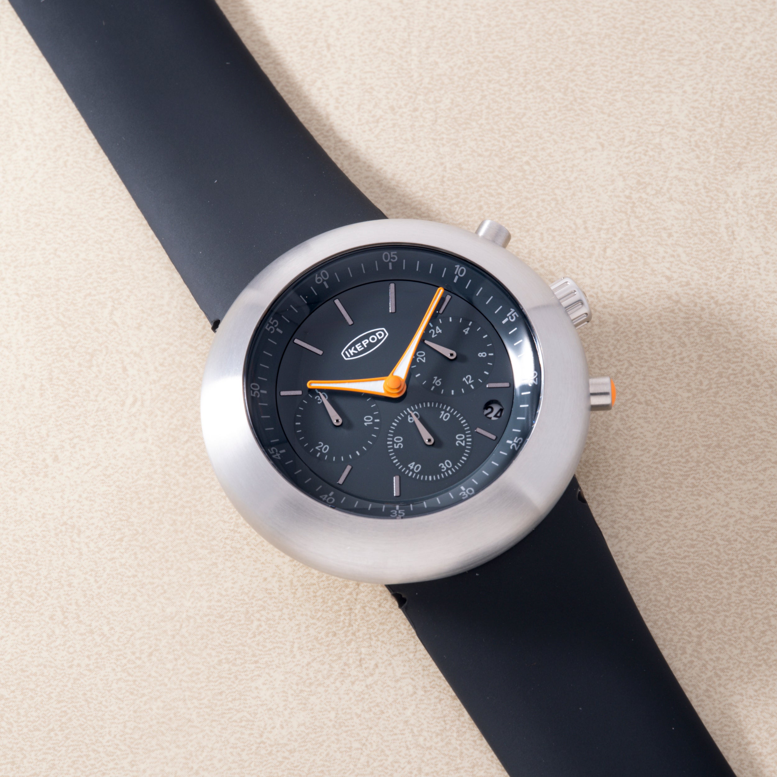 White chronograph watch with black dial, orange hands, and black rubber strap on beige background.