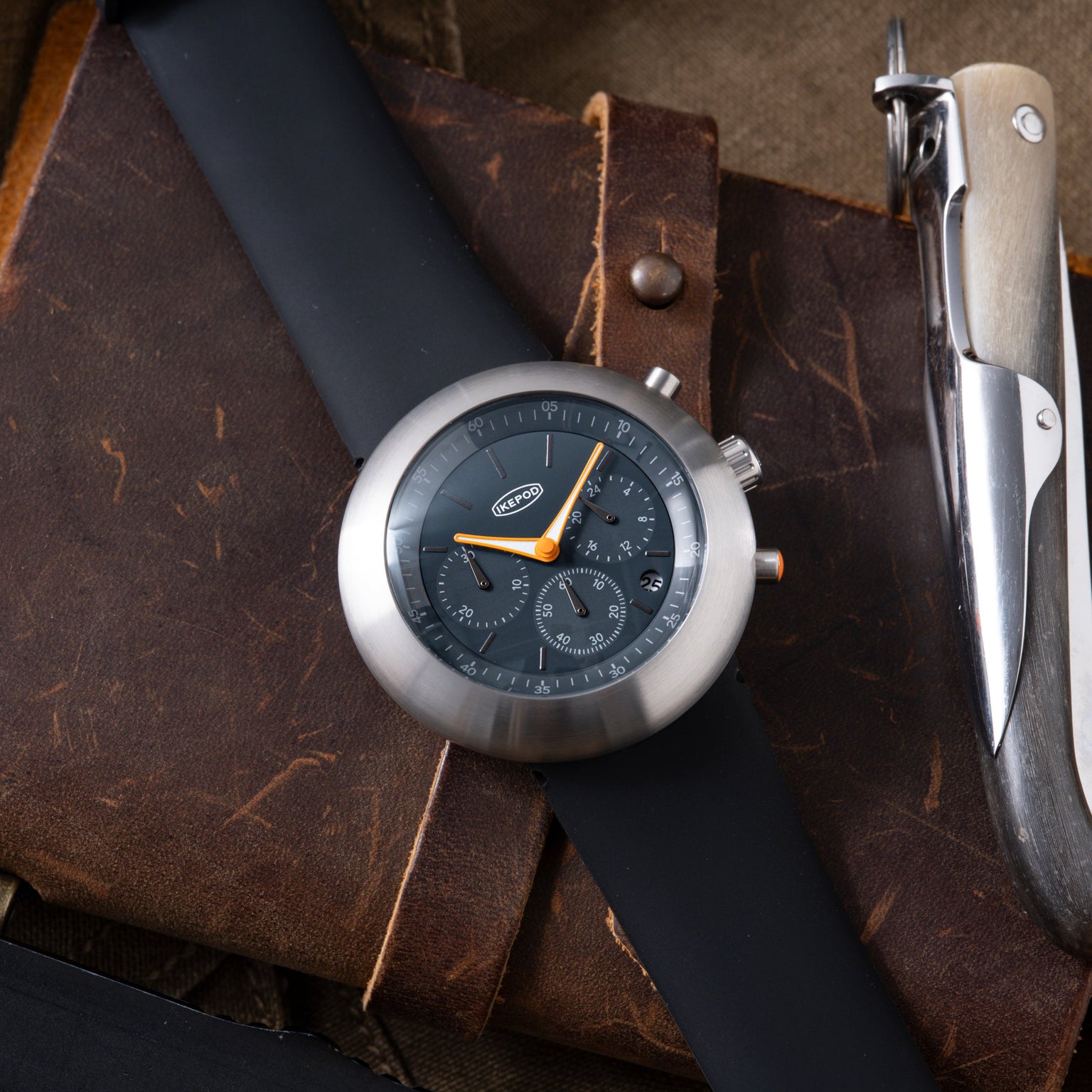 Ikepod Chronopod chronograph watch with dark dial and orange hands on brown leather strap, displayed with multi-tool.