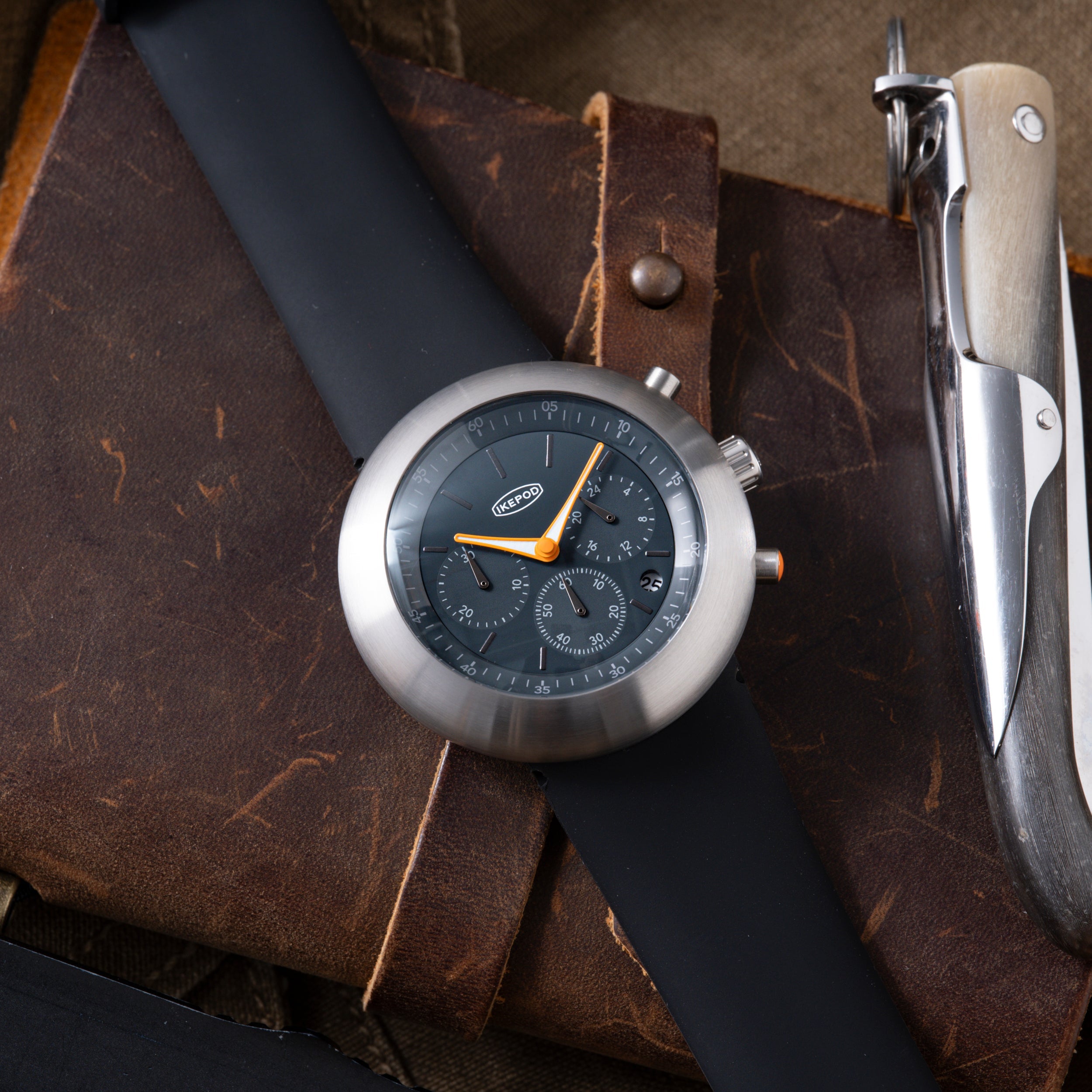 Ikepod Chronopod chronograph watch with dark dial and orange hands on brown leather strap, displayed with multi-tool.