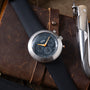 Ikepod Chronopod chronograph watch with dark dial and orange hands on brown leather strap, displayed with multi-tool.
