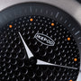 Black watch dial with textured dot pattern, white hands, orange accent markers, and Ikepod branding.