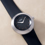 Round watch with silver case, black dial featuring orange dot markers, white hands, and black rubber strap.