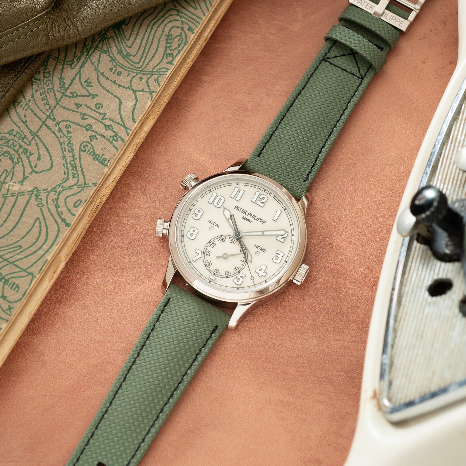 Silver-tone watch with cream dial, Arabic numerals, and green fabric strap on arm with map and book nearby.
