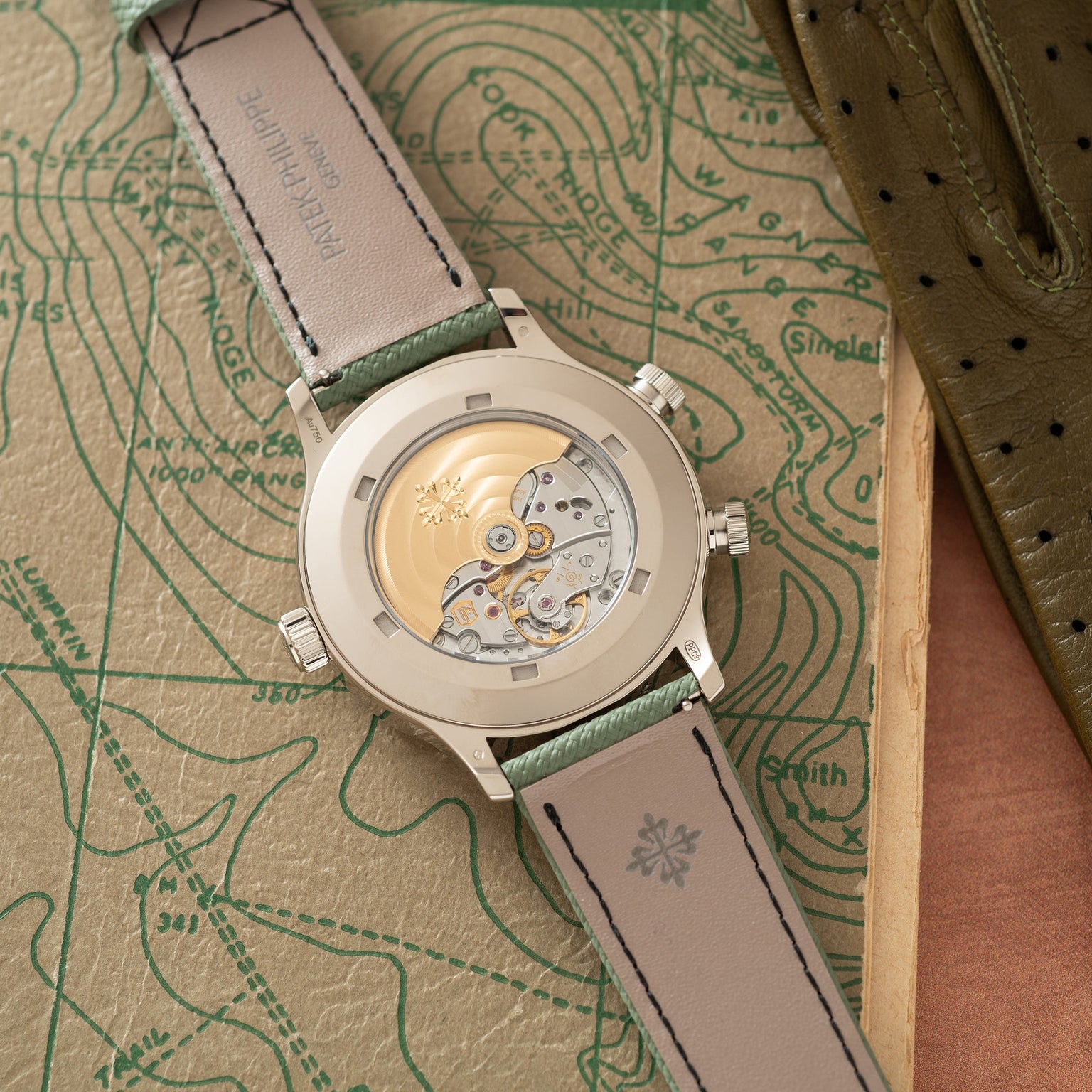 Silver-tone watch with visible skeleton movement dial and gray-green leather strap on topographic map background.