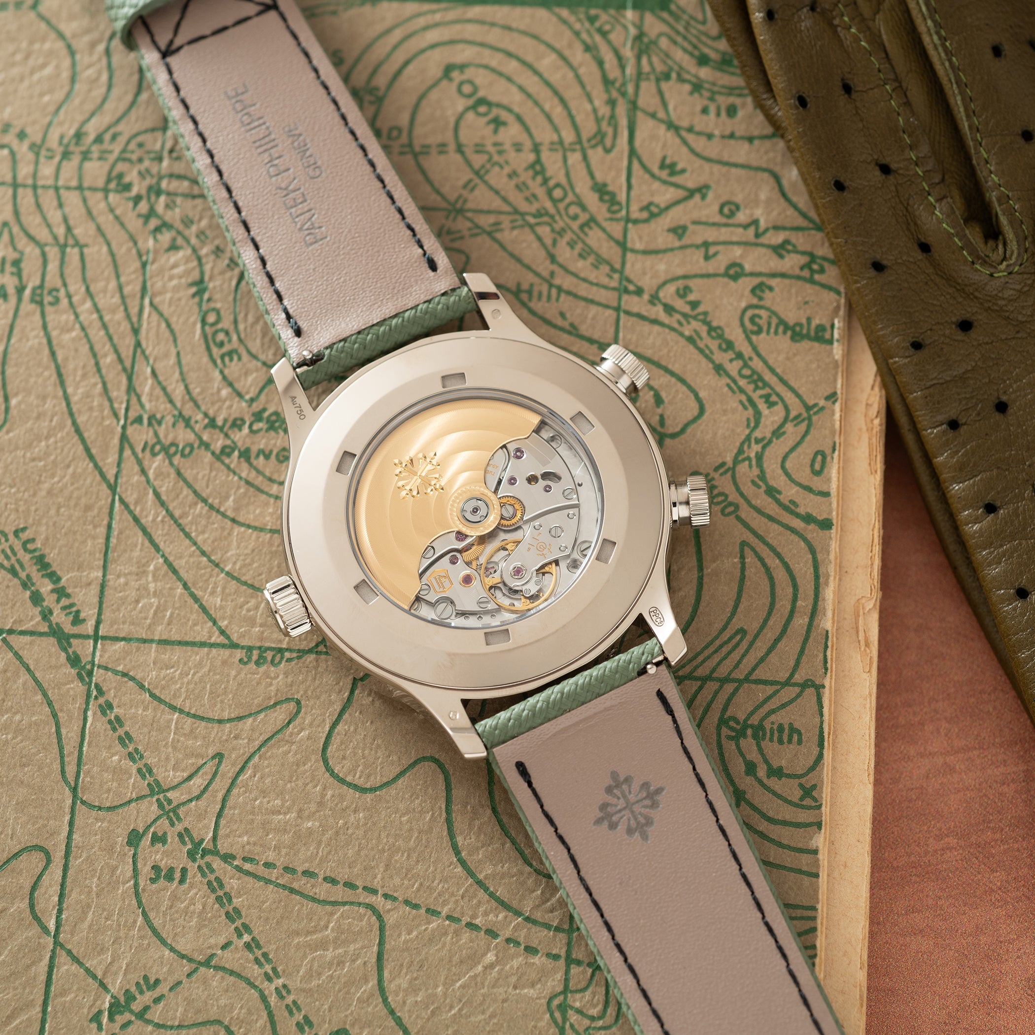 Silver-tone watch with visible skeleton movement dial and gray-green leather strap on topographic map background.
