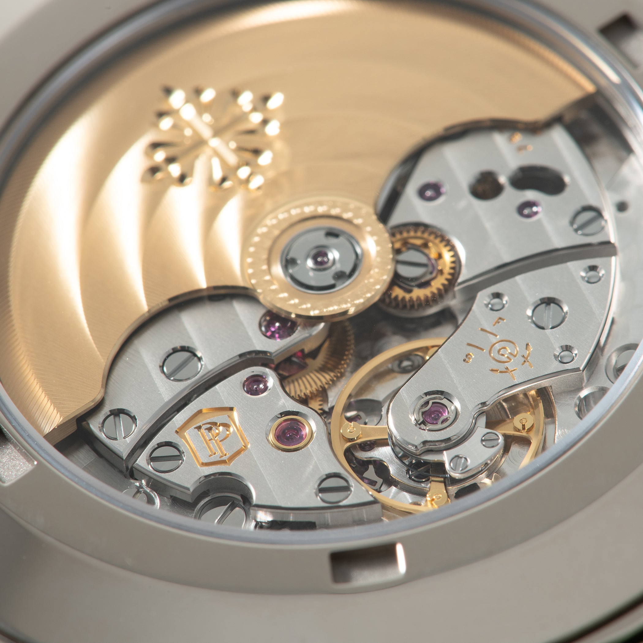 Close-up of exposed watch movement with polished steel plates, golden gears, ruby jewels, and engraved Patek Philippe logo.