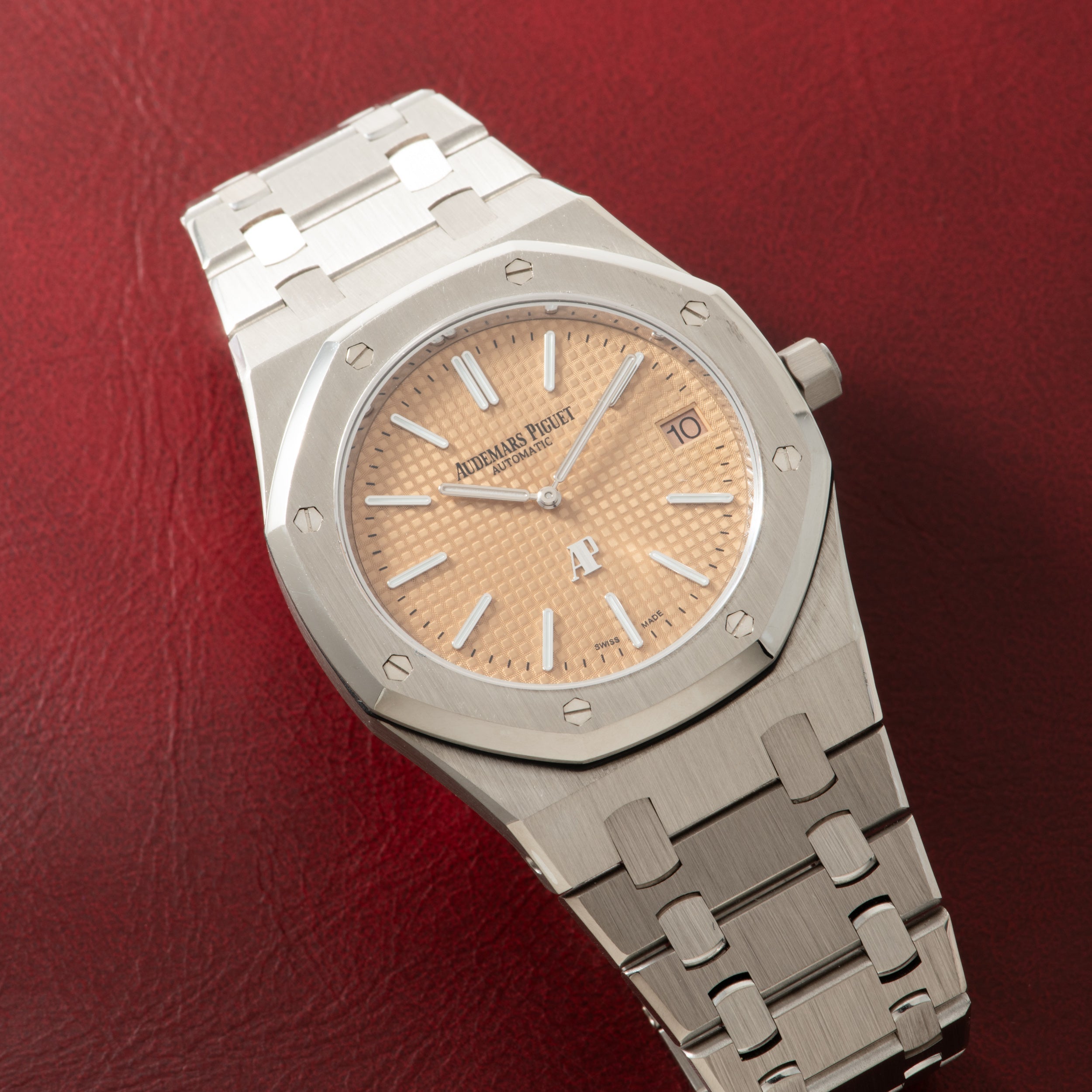 Stainless steel watch with salmon-colored dial, white applied indices, and matching metal bracelet on red background.