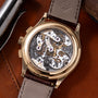 Gold-tone chronograph watch with exposed skeletal movement, white and black dial components, gray leather strap on brown back