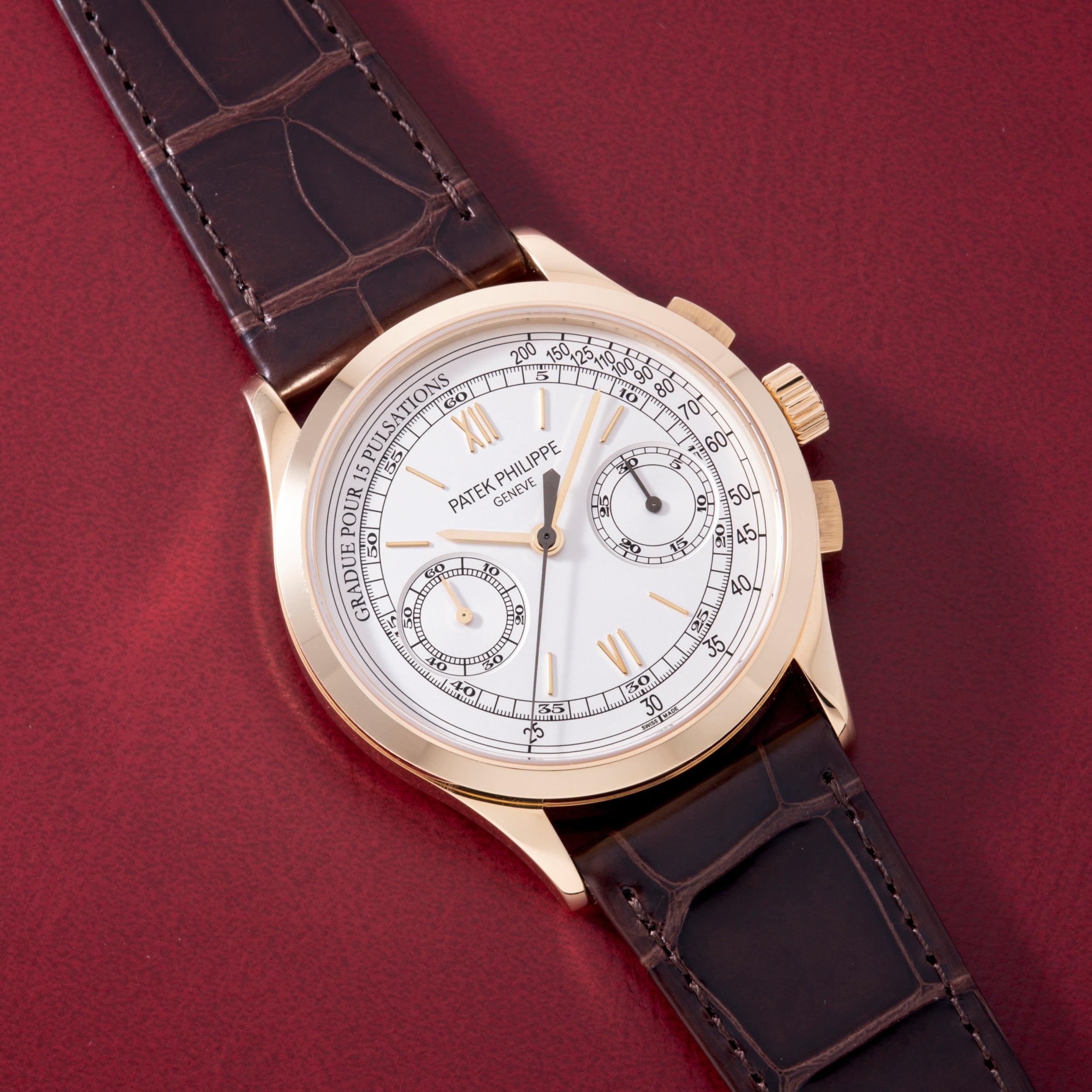 Gold-tone chronograph watch with white dial, two subdials, Roman numerals, and dark crocodile leather strap on burgundy backg