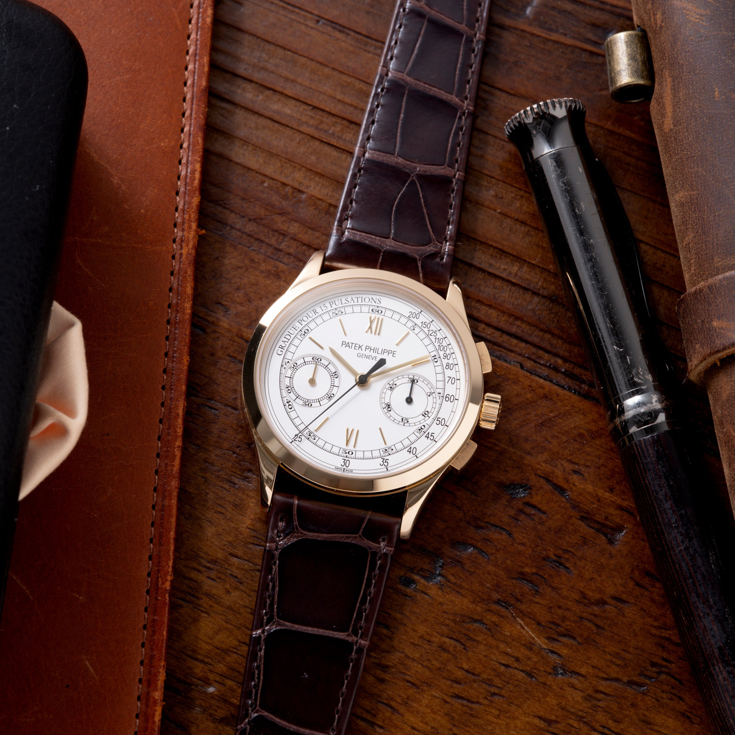 Patek Philippe chronograph with white dial, two subdials, and dark crocodile leather strap on wooden surface.