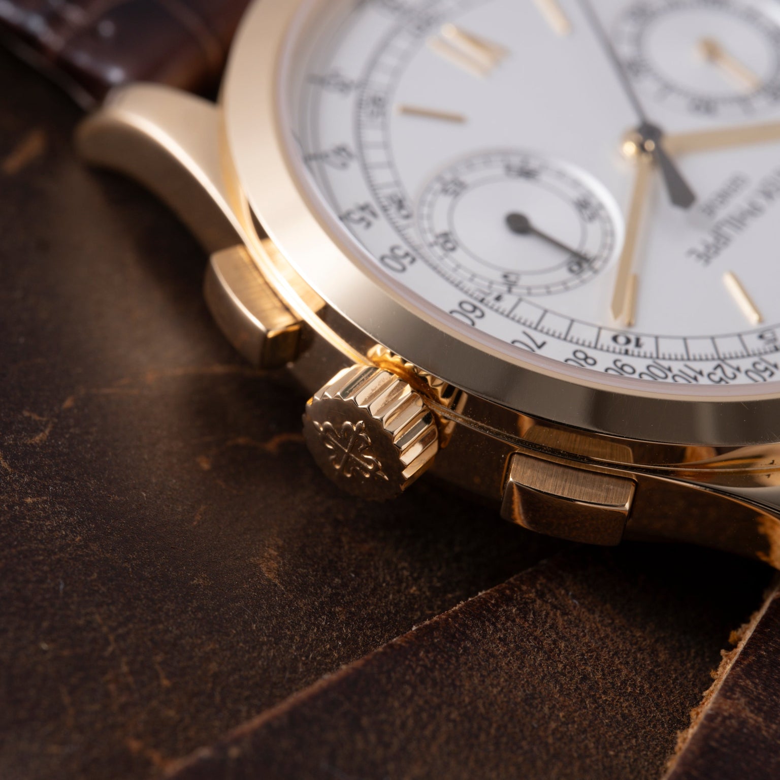 Gold chronograph watch with white dial, subsidiary dials, and textured crown on dark leather surface.