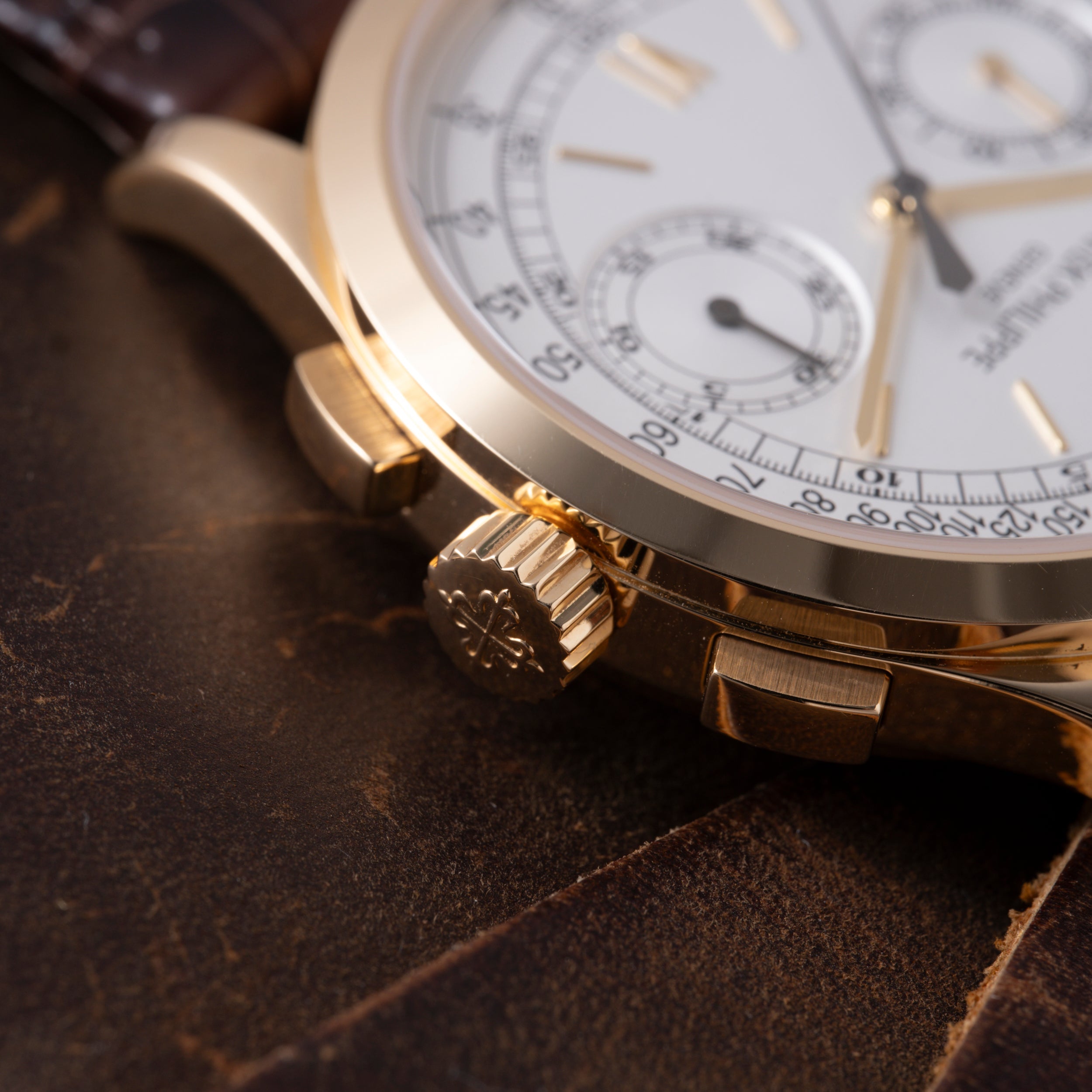 Gold chronograph watch with white dial, subsidiary dials, and textured crown on dark leather surface.