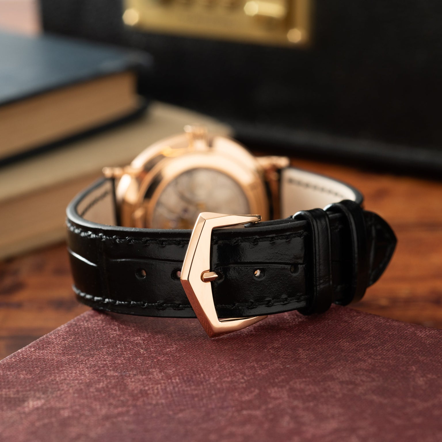 Gold-tone watch with black leather strap and rose gold buckle displayed on burgundy fabric.