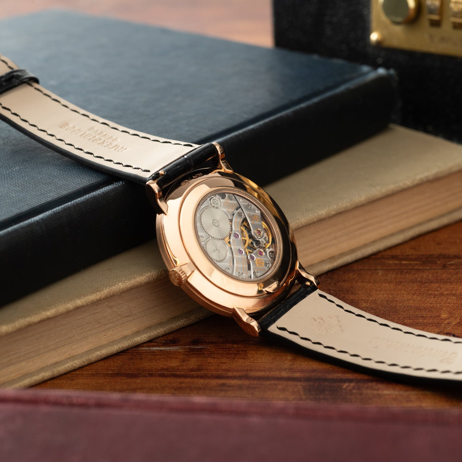 Rose gold watch with skeletonized dial and black leather strap with white stitching, displayed on books.