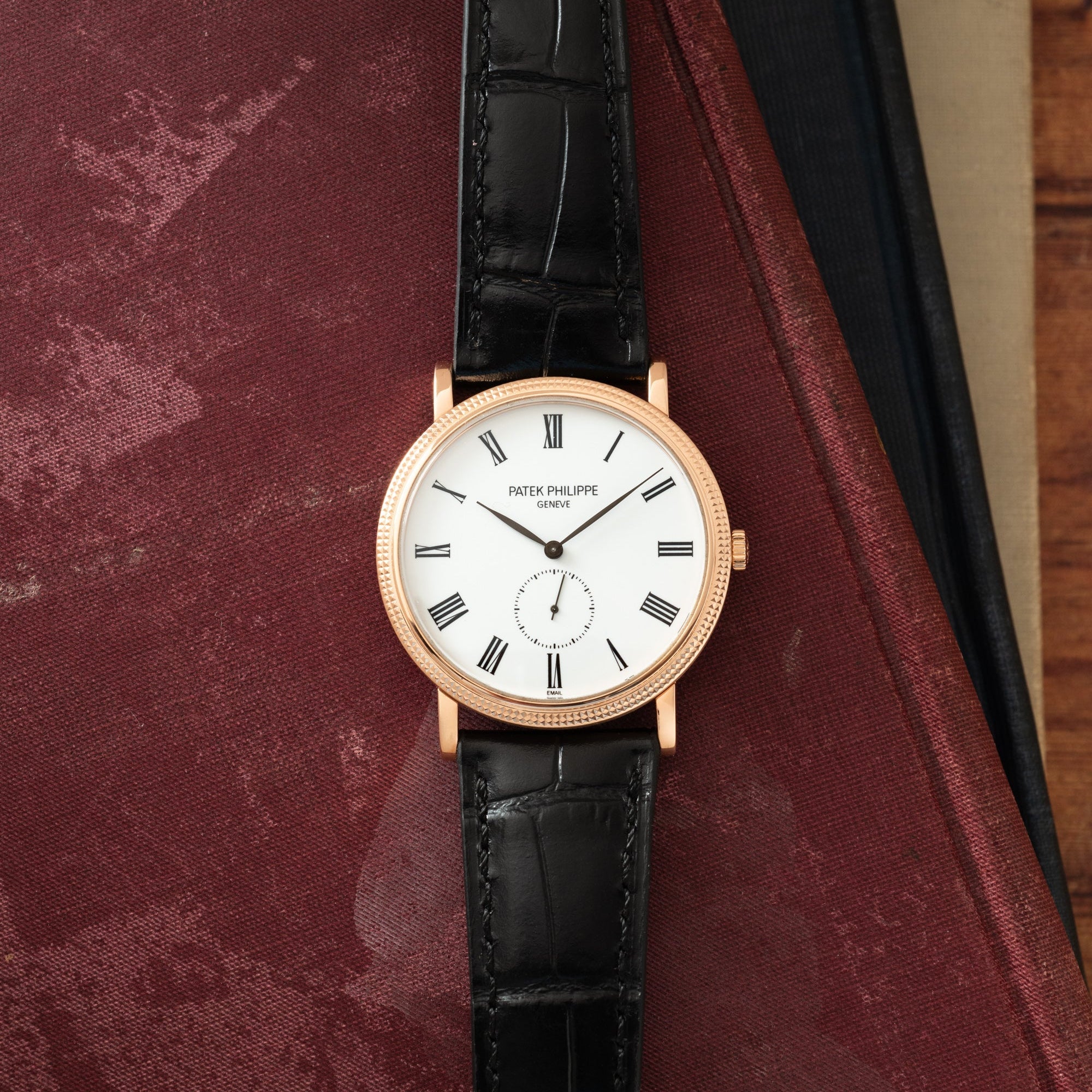Rose gold round watch with white dial, Roman numerals, black leather strap, displayed on burgundy fabric.