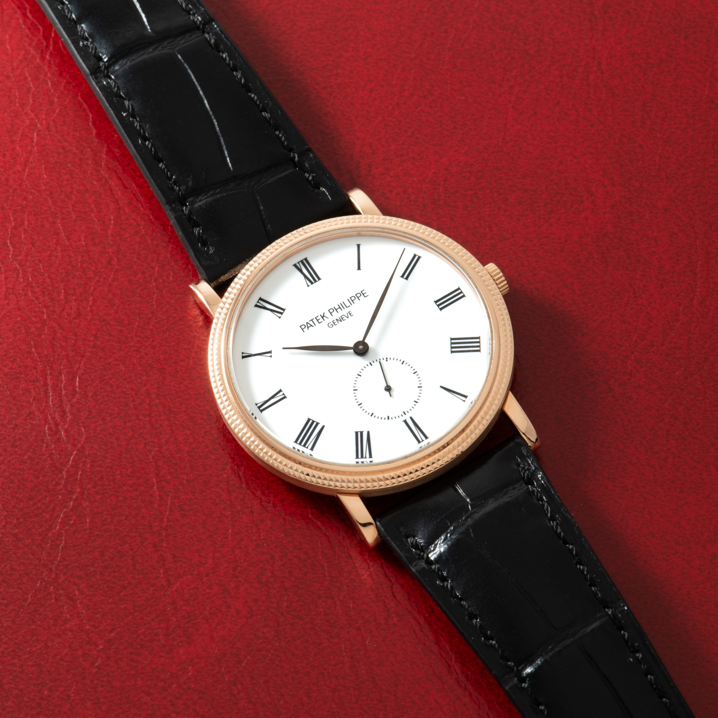 Gold-tone watch with white dial, Roman numerals, subdial, and black crocodile leather strap on red background.