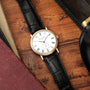 Gold-tone watch with white dial, Roman numerals, and black crocodile leather strap on wooden surface.