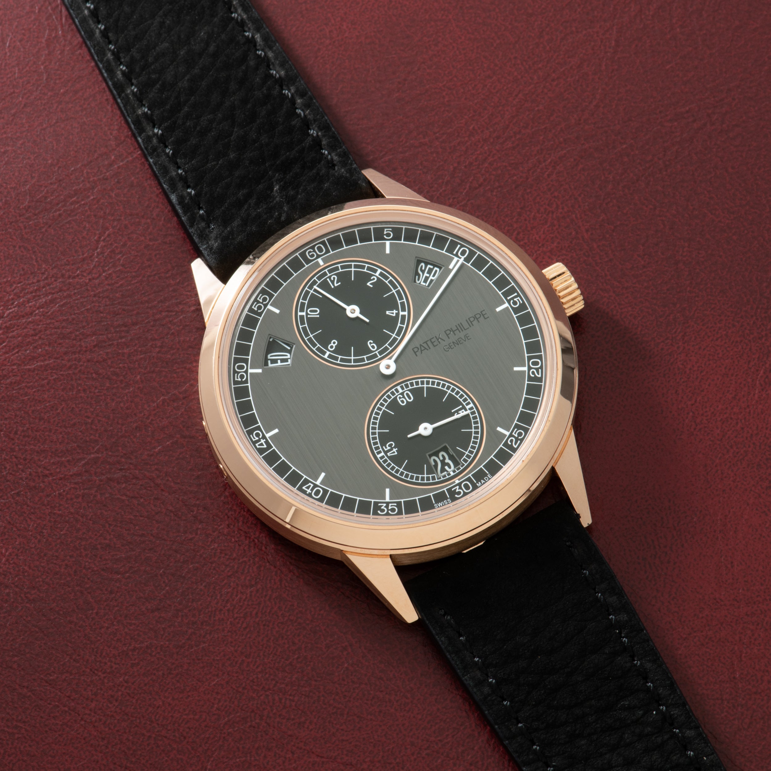 Rose gold watch with gray dial, two subdials, and black leather strap on burgundy background.