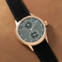 Rose gold watch with gray dial, two subdials, and black leather strap on tan background.