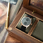 Silver-tone watch with dual subdials and olive green fabric strap displayed in wooden box with tan interior and brass hardwar