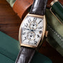 Gold-tone rectangular watch with white dial, dual subdials, Arabic numerals, black leather strap on colored fabric background