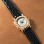 Gold-toned watch with cream dial, Roman numerals, and black crocodile leather strap.
