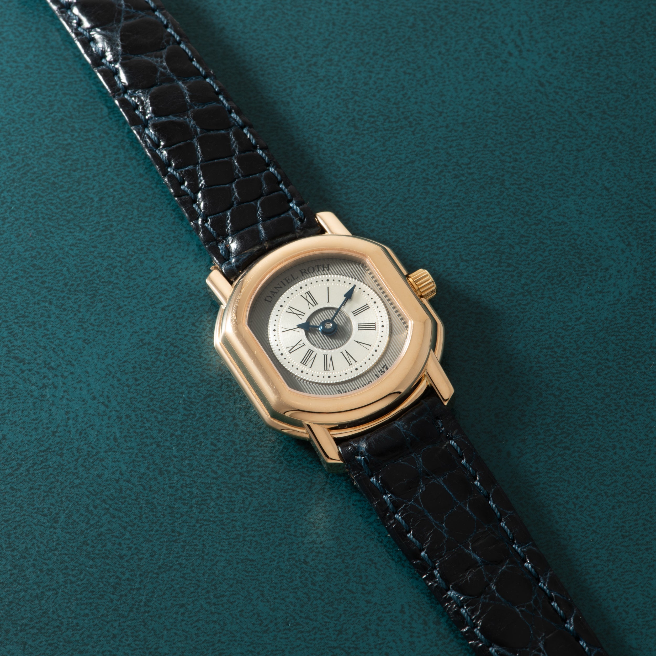 Gold-tone watch with cream dial, Roman numerals, and black crocodile leather strap on teal background.