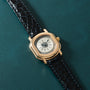 Gold-tone watch with cream dial, Roman numerals, and black crocodile leather strap on teal background.
