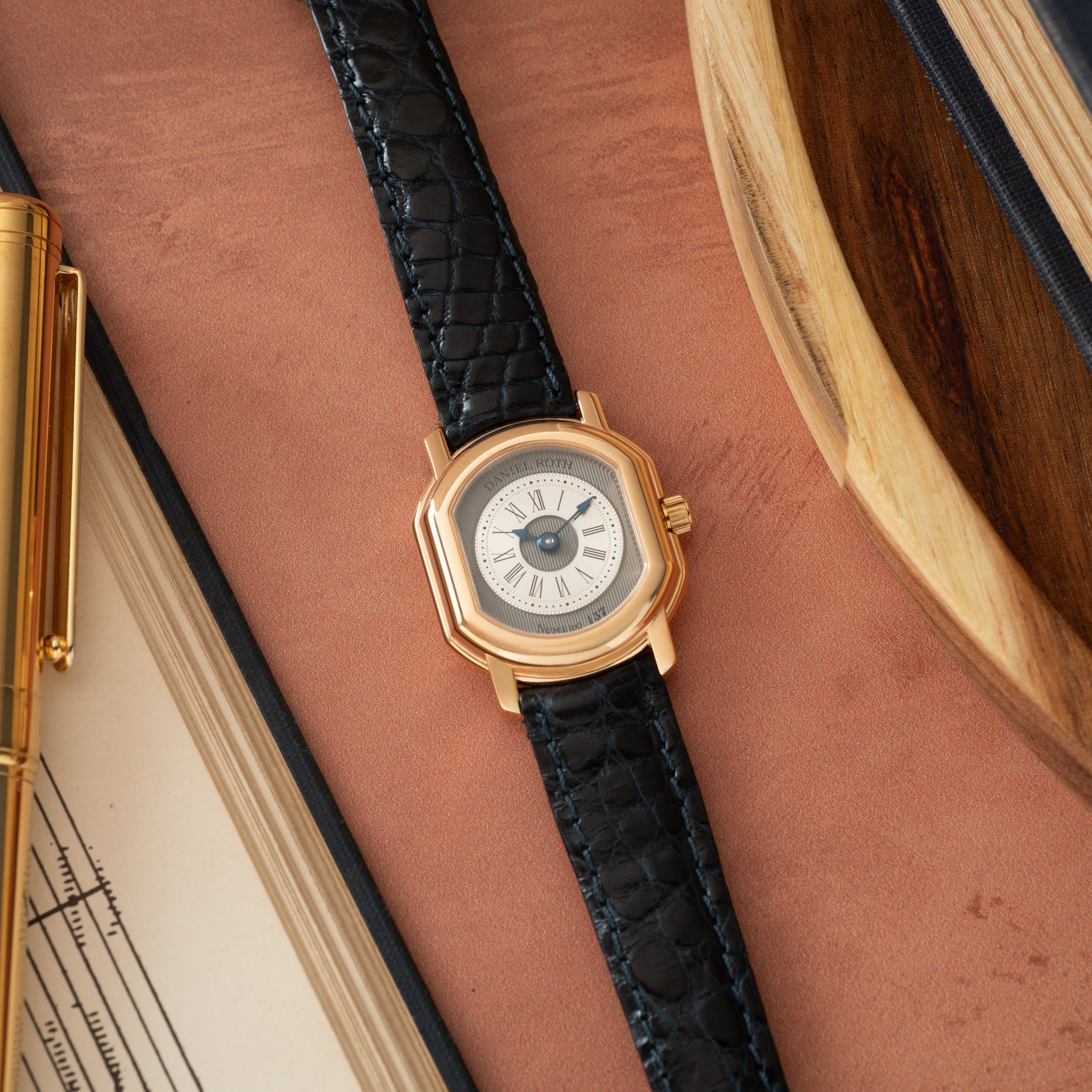 Gold-tone watch with white dial, Roman numerals, and black crocodile leather strap on terracotta surface.