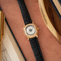 Gold-tone watch with white dial, Roman numerals, and black crocodile leather strap on terracotta surface.