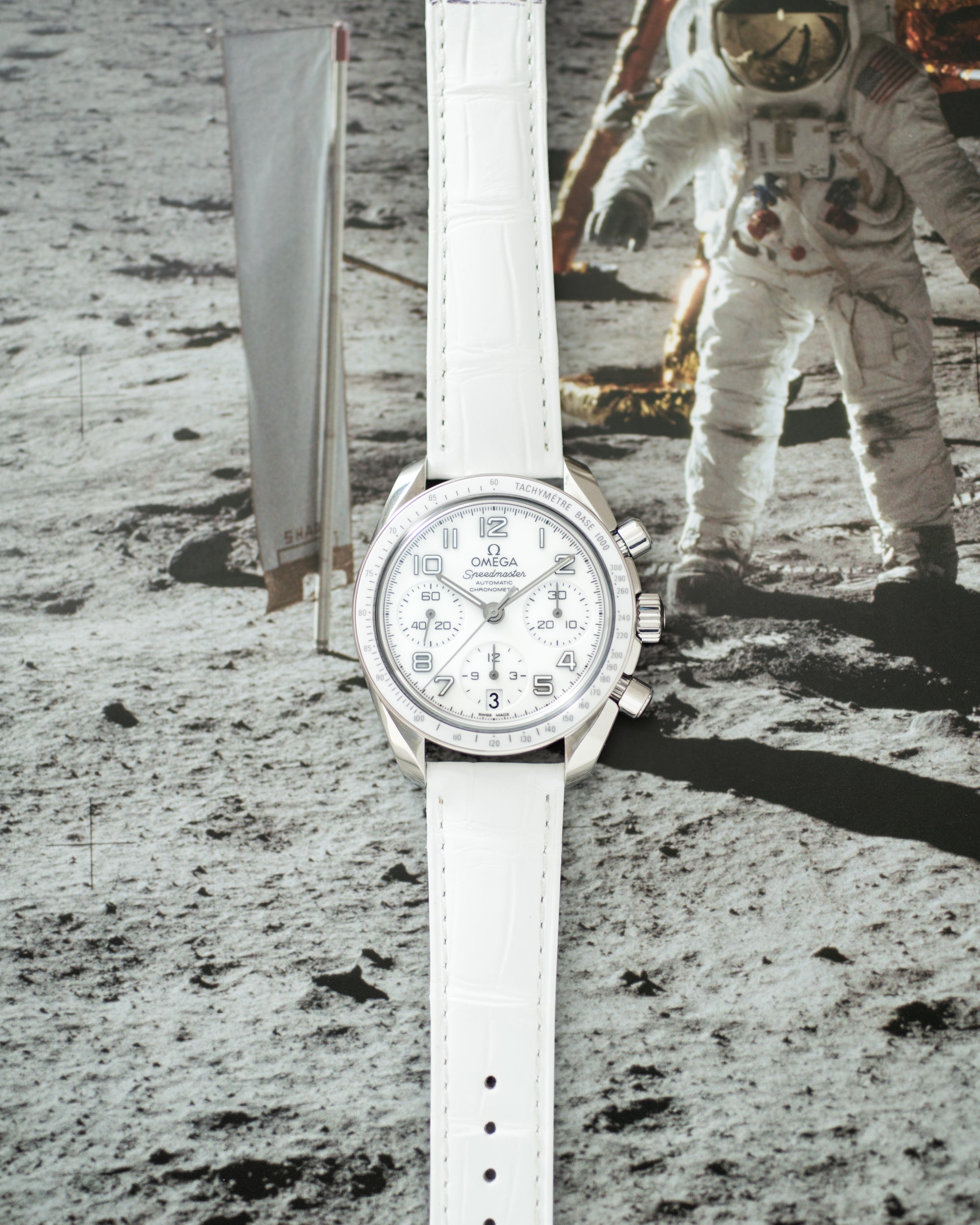 Silver chronograph watch with white dial and white leather strap on lunar surface background.