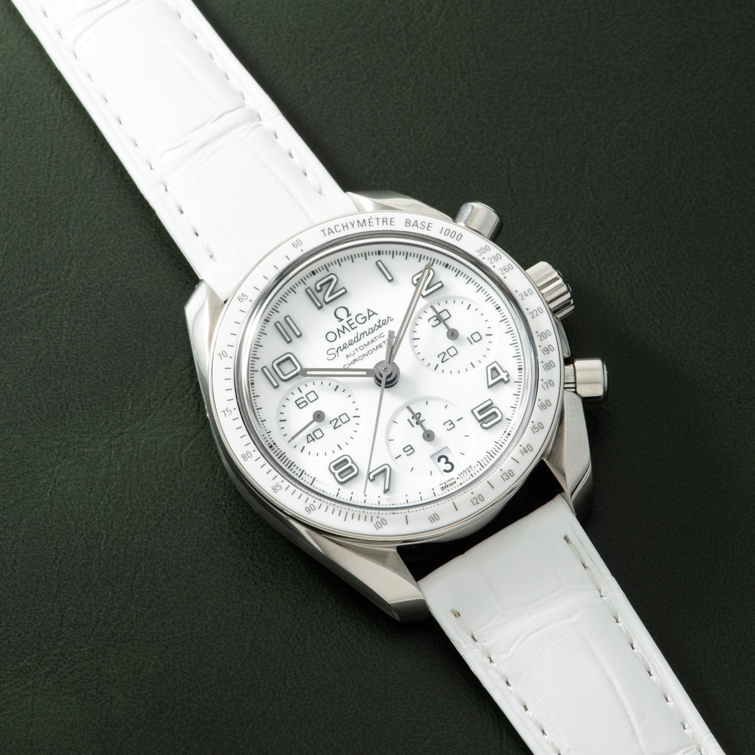 Omega Speedmaster chronograph with white dial, tachymetre bezel, and white leather strap on green background.