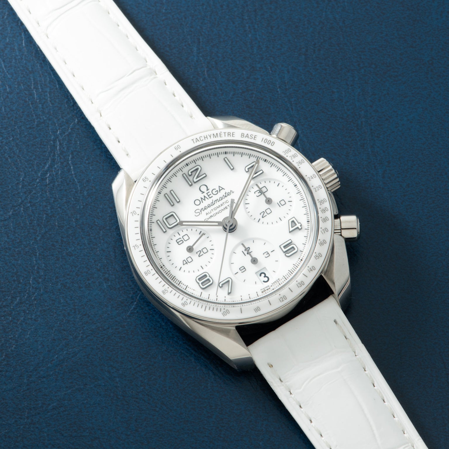 Silver chronograph watch with white dial, Arabic numerals, tachymeter bezel, and white leather strap on blue background.