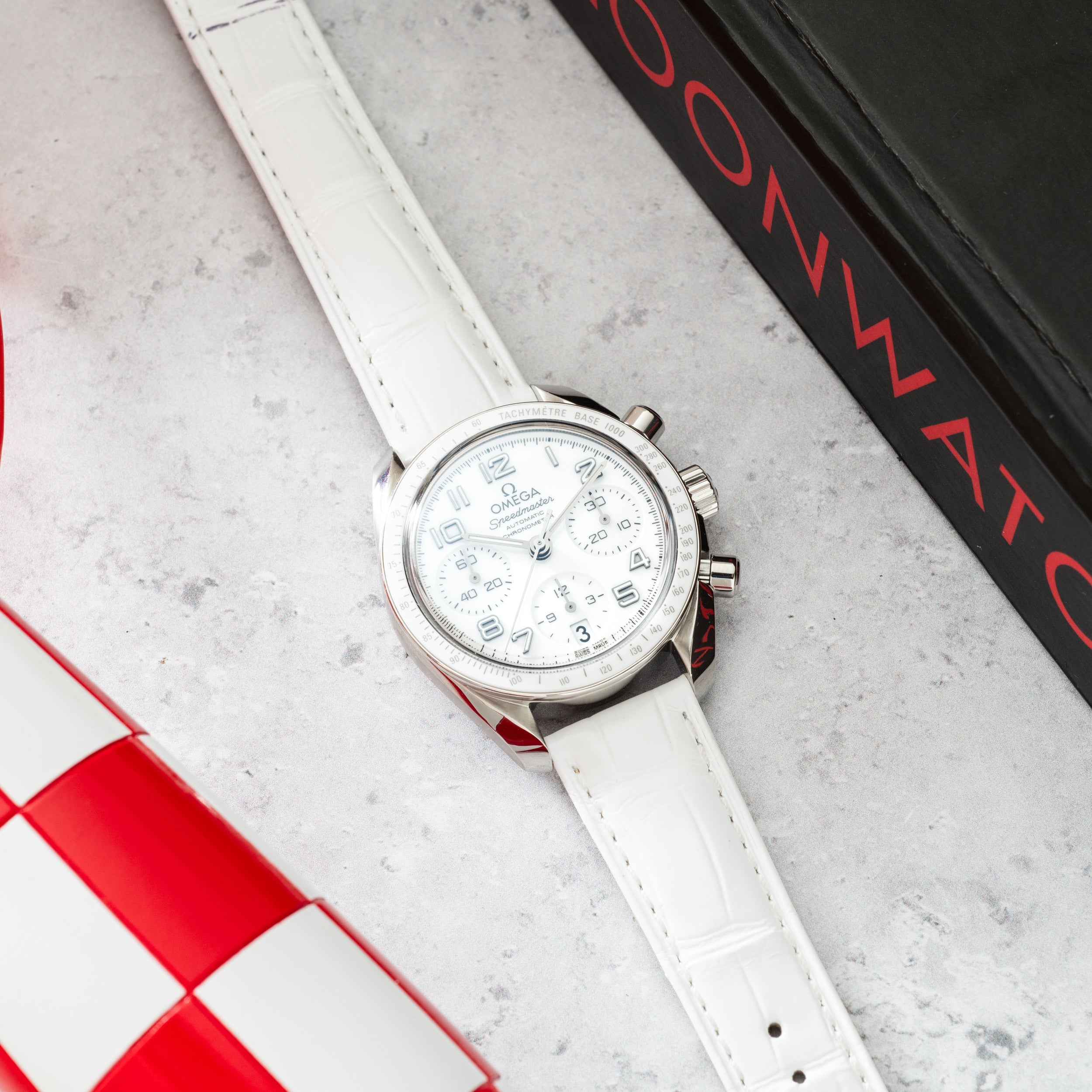 Silver-tone chronograph watch with white dial and white leather strap on concrete surface with red and black branding.