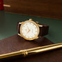 Gold-tone watch with white dial, gold hour markers, brown leather strap, displayed on brown textured surface with pen.