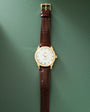 Gold-tone watch with white dial, hour markers, and burgundy crocodile-embossed leather strap with gold buckle.