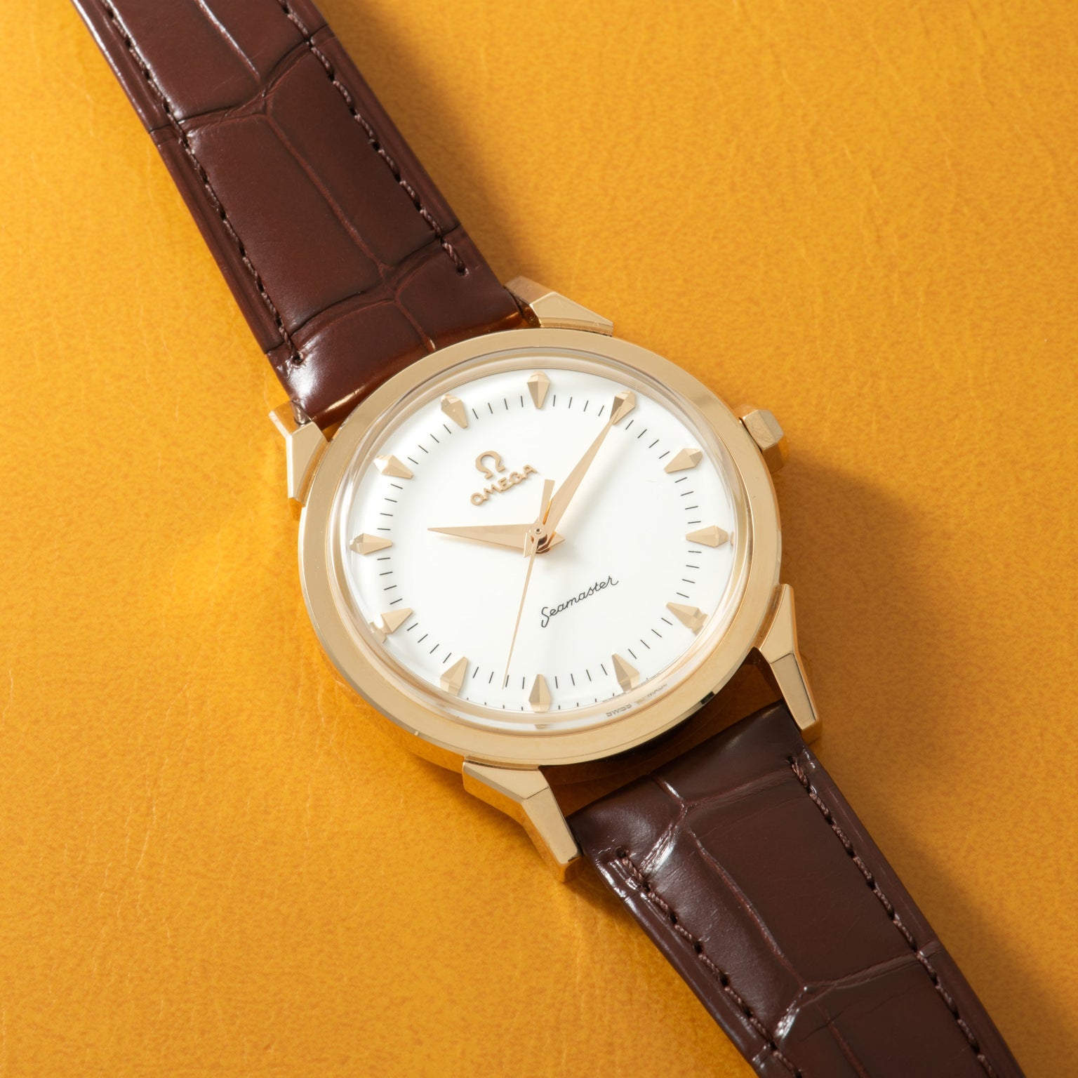 Gold-tone watch with white dial, gold hour markers, and dark brown crocodile-textured leather strap on yellow background.